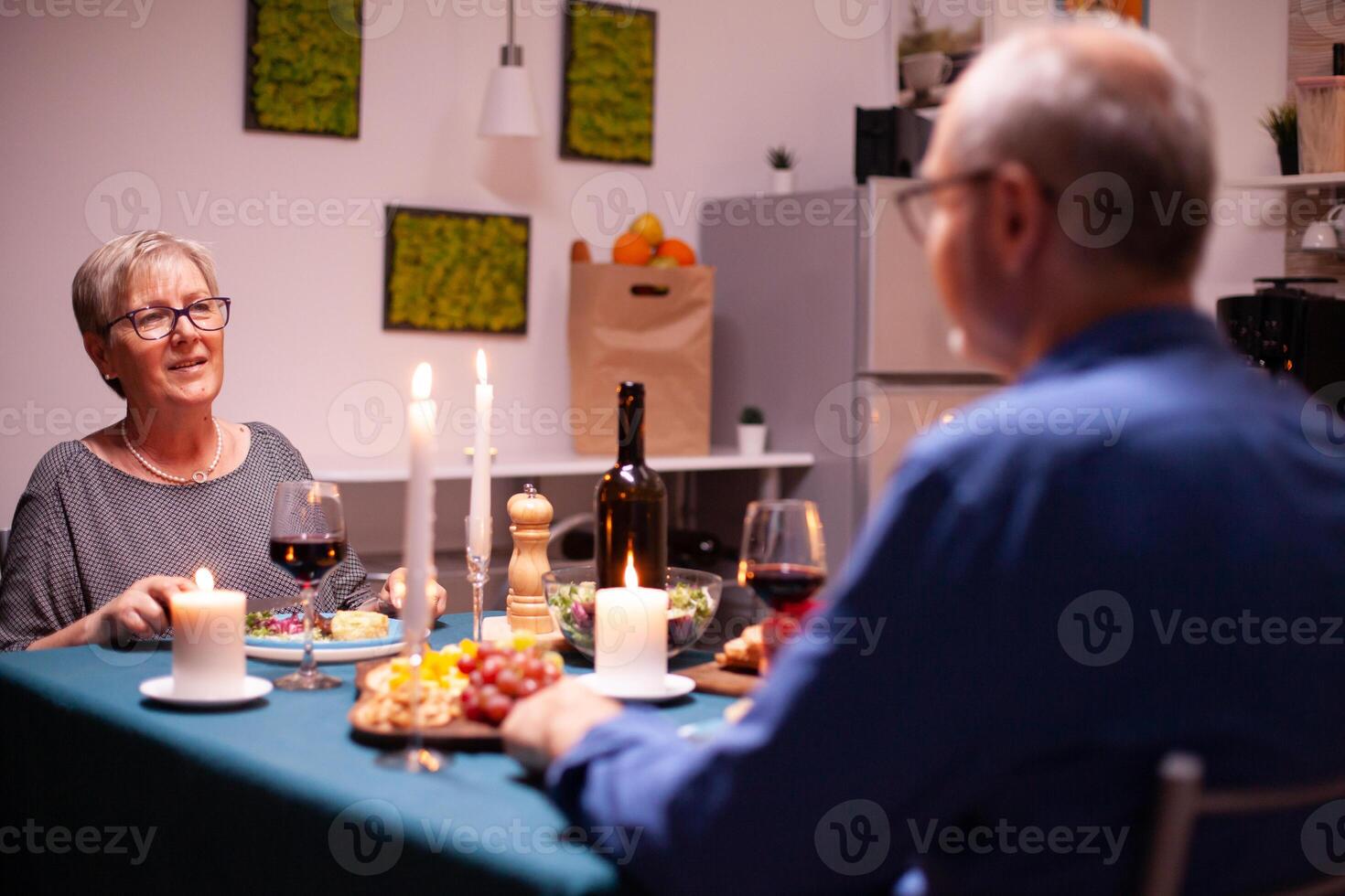 Mature woman having a conversation with husband while holding glass of red wine in kitchen. Senior couple sitting at the table in kitchen, talking, enjoying the meal, celebrating their anniversary in the dining room. photo