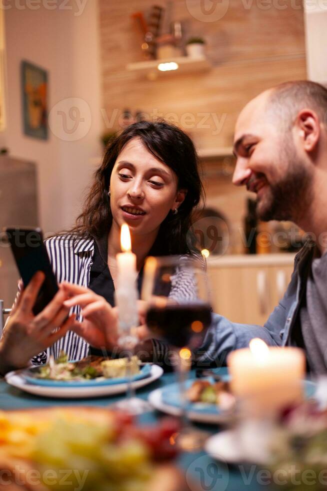 Wife showing husband photos on smartphone during festive relationship anniversary. Adults sitting at the table in the kitchen browsing, searching, using smartphones, internet, celebrating