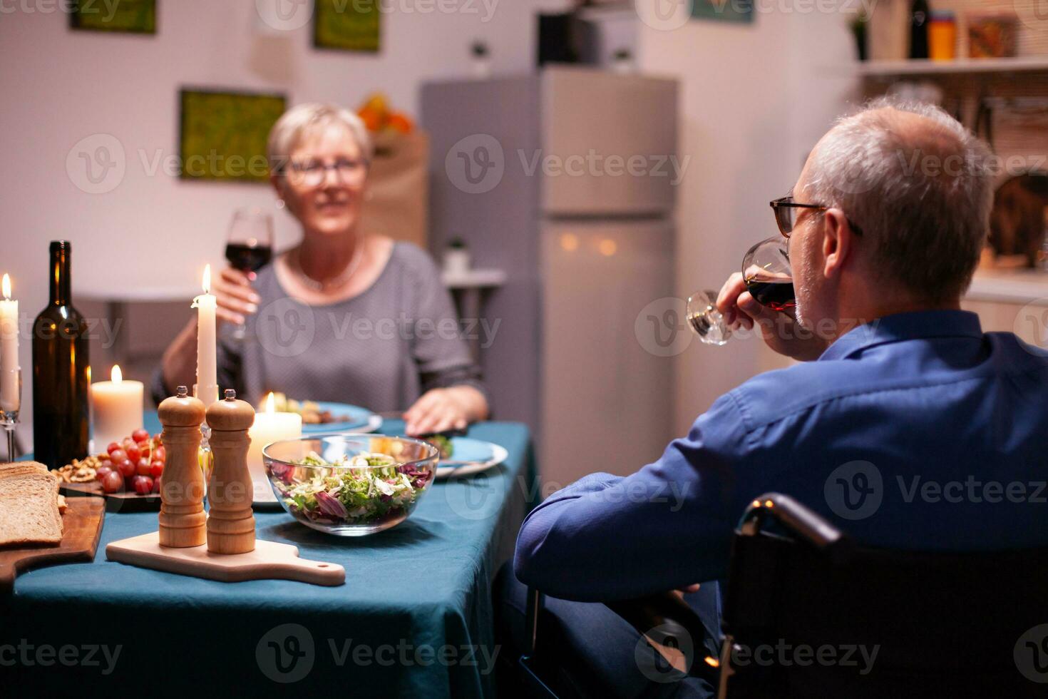 paralizado hombre en silla de ruedas Bebiendo vino durante cena con esposa. contento alegre mayor mayor Pareja comida juntos en el acogedor cocina, disfrutando el comida, celebrando su aniversario. foto