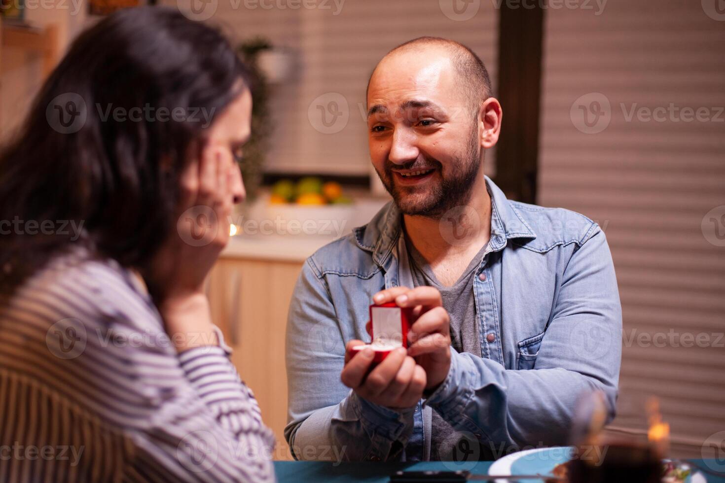 Young man asking wife to marry him during dinner in kitchen. Man making proposal to his girlfriend in the kitchen during romantic dinner. Happy caucasian woman smiling being speechless photo