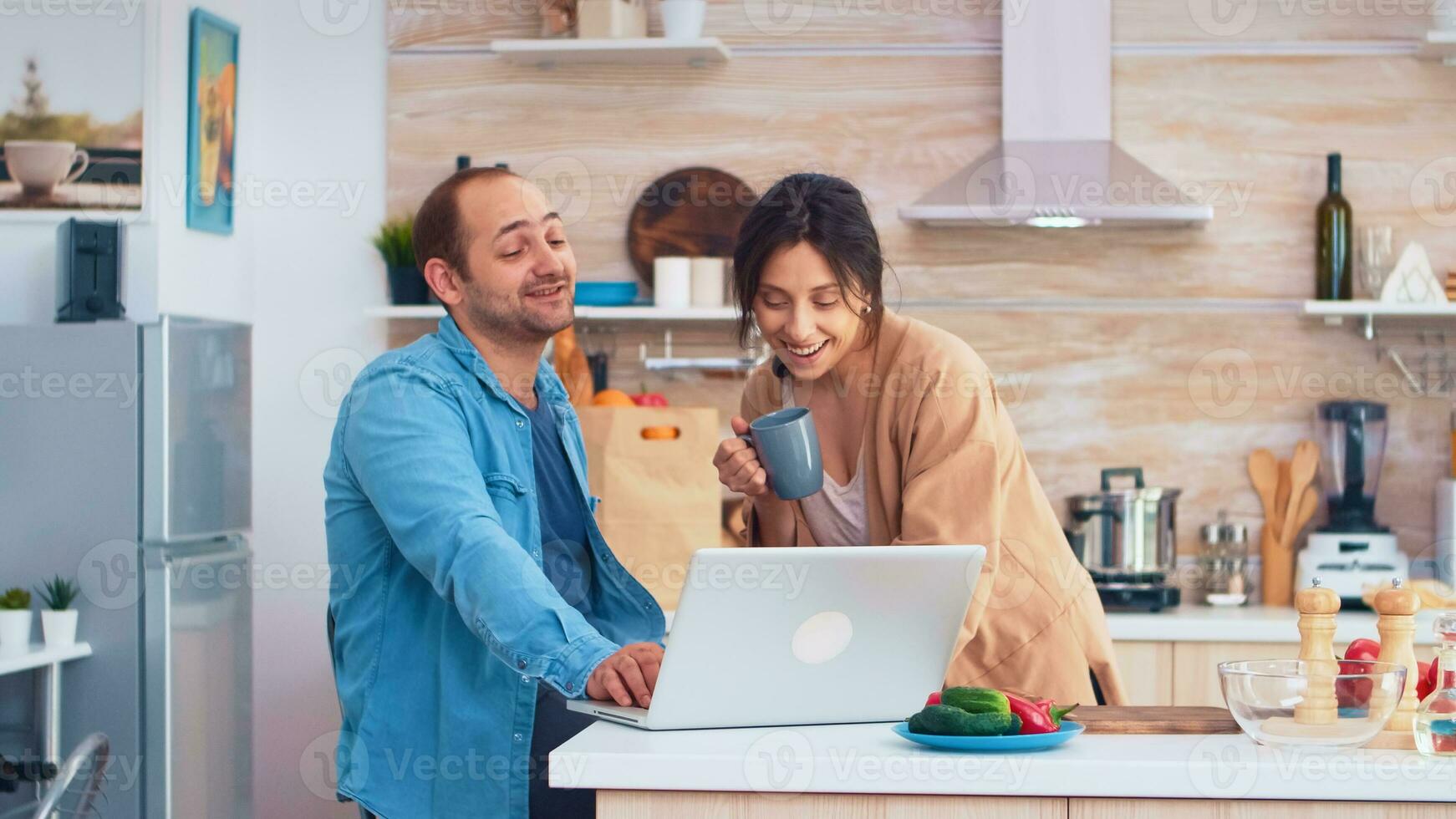 Entrepreneur working on laptop in kitchen and wife holding a cup of coffee looking at his work. Husband and wife cooking recipe food. Happy healthy together lifestyle. Family searching for online meal. Health fresh salad photo