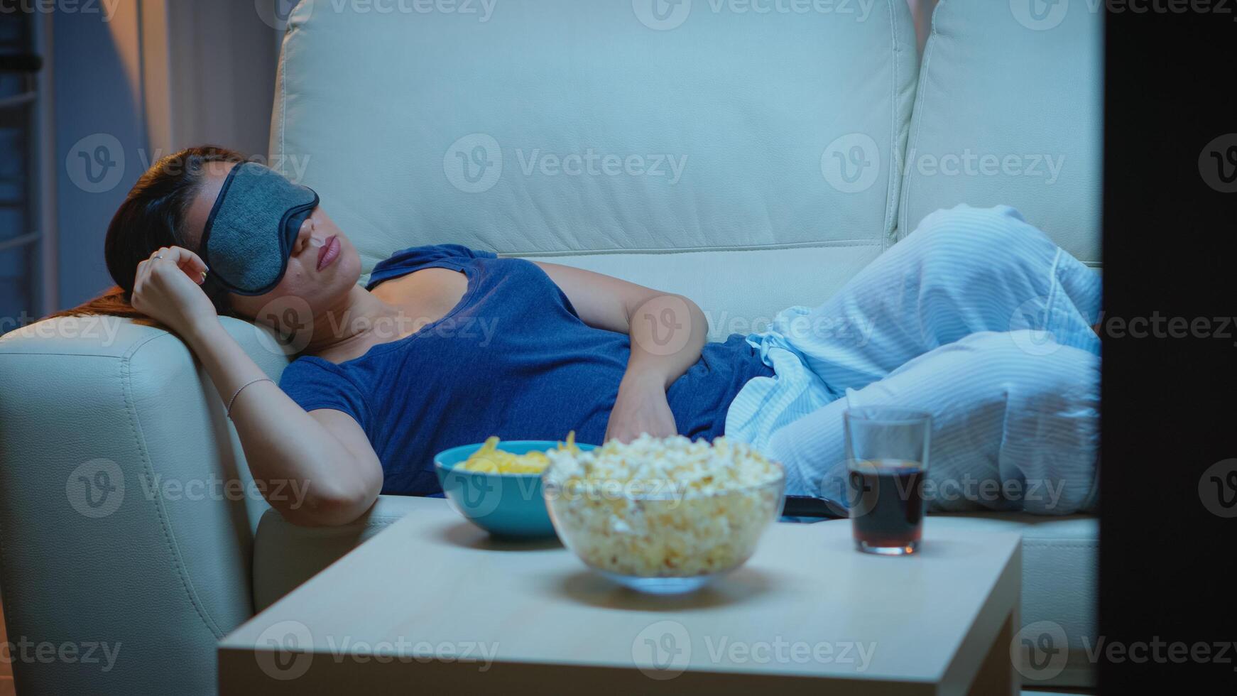Woman sleeping with eye covering mask in front of tv lying on couch. Tired exhausted lonely sleepy lady in pajamas falling asleep on sofa in front television, closing eyes while watching movie. photo