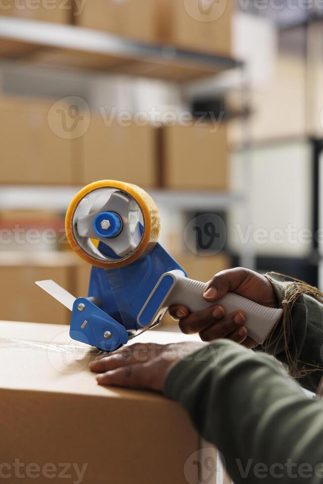 Cheerful employee using adhesive tape to pack cardboard box, preparing clients packages for delivery in warehouse. Storage room supervisor wearing industrial overall working at online orders. Close up photo