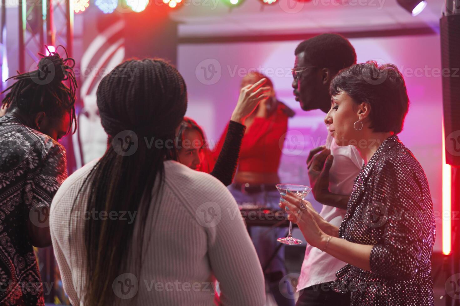 Young friends drinking alcohol and partying on crowded club dancefloor illuminated with spotlights. Diverse people dancing and enjoying nightlife activity at nightclub discotheque photo