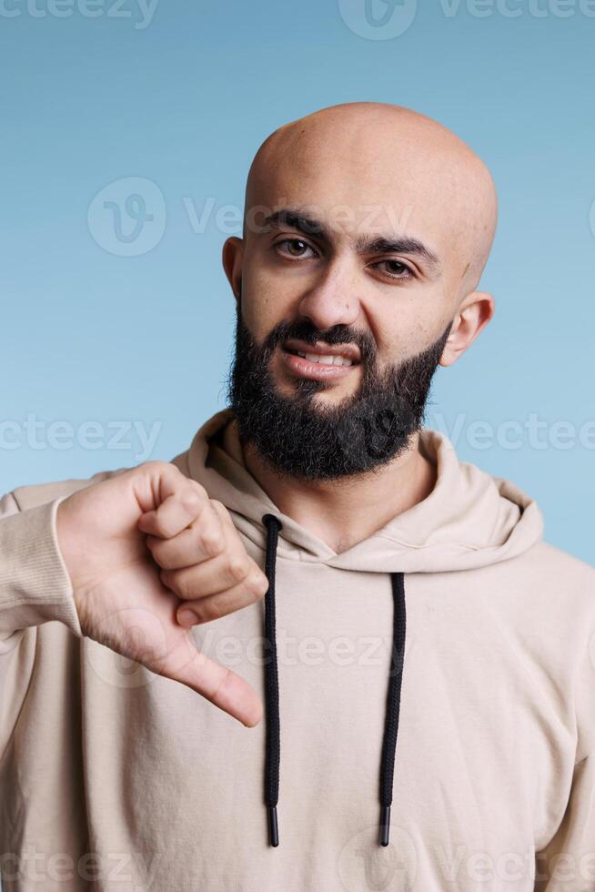 Arab man looking at camera with thumb down gesture to express negative feelings. Young person wearing casual hoodie clothes showing dislike sign with right hand studio portrait photo