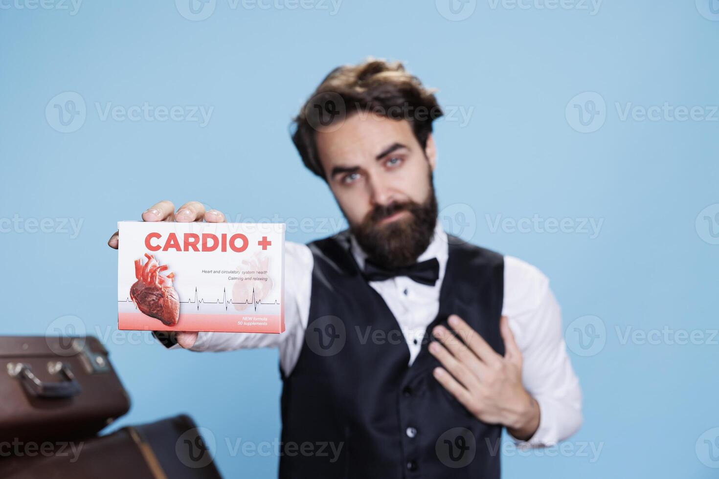 Hotel porter with heart problems presents drugs on camera, holding box of medicaments that cure cardiovascular disease. Elegant bellboy shows medication in studio, suit and tie. photo