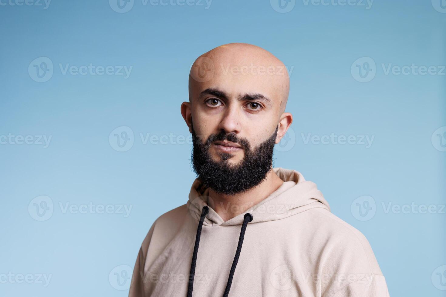 Young bald arab man with relaxed facial expression studio portrait. Bearded arabian person with neutral emotions wearing casual beige hoodie and looking at camera on blue background photo
