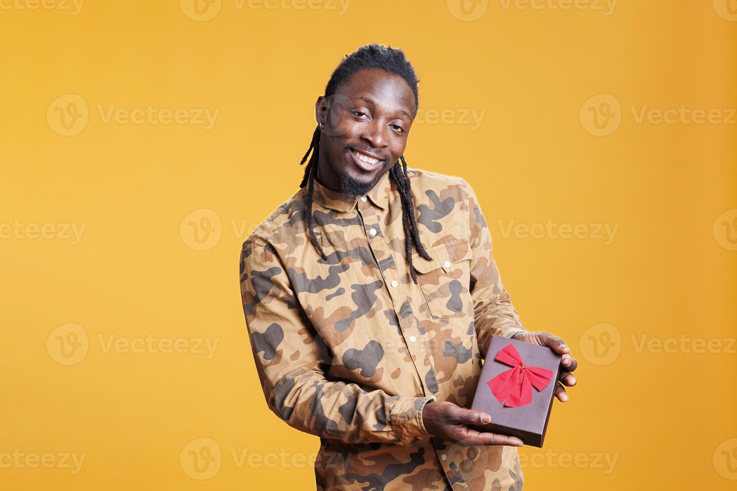 Confident smiling man presenting wrapped present box for girlfriend, posing for valentine s day in studio over yellow background. Handsome african american person celebrating love holiday. photo
