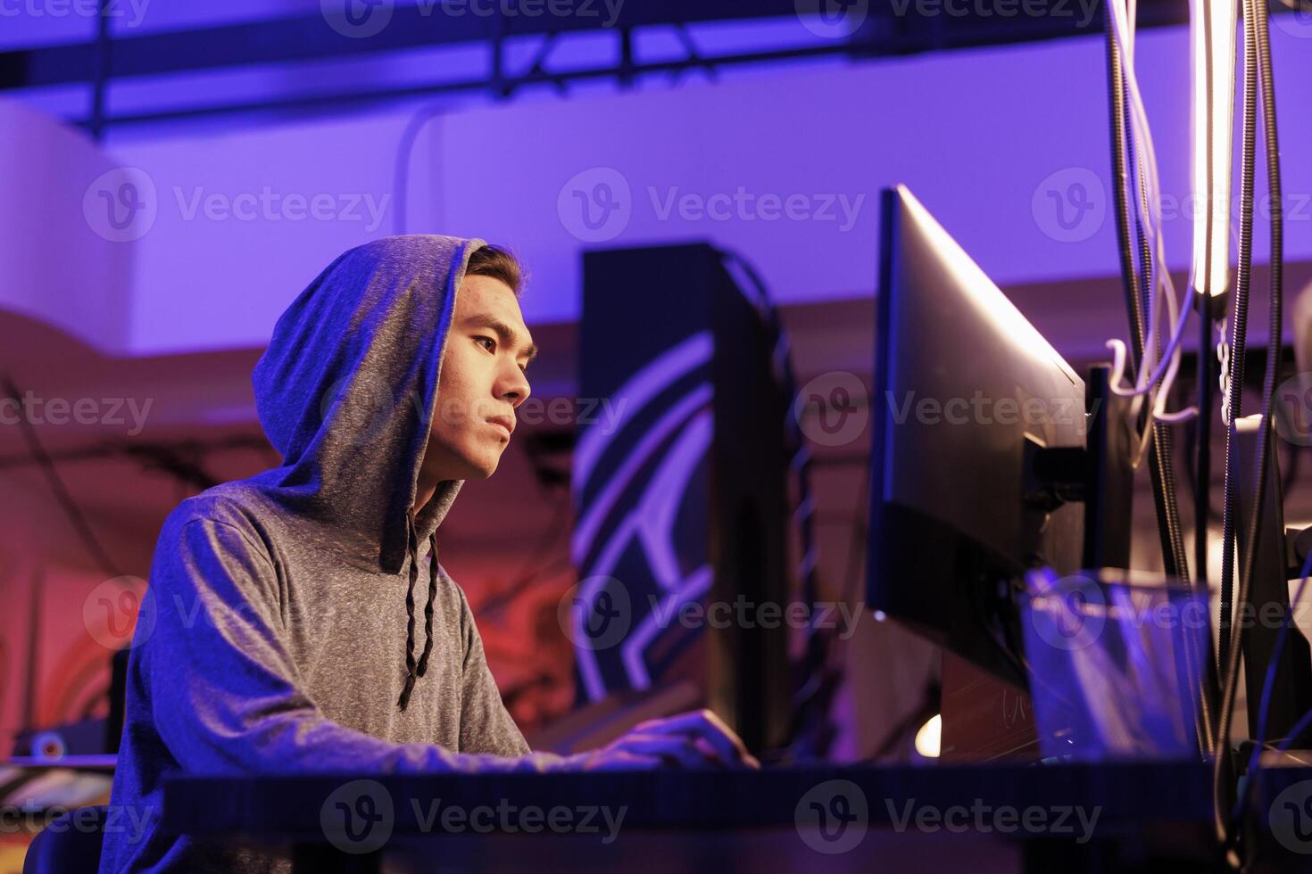 Asian hacker breaking law while doing data mining using malicious software. Young man in hood hacking into computer system while cracking password in abandoned warehouse at night photo