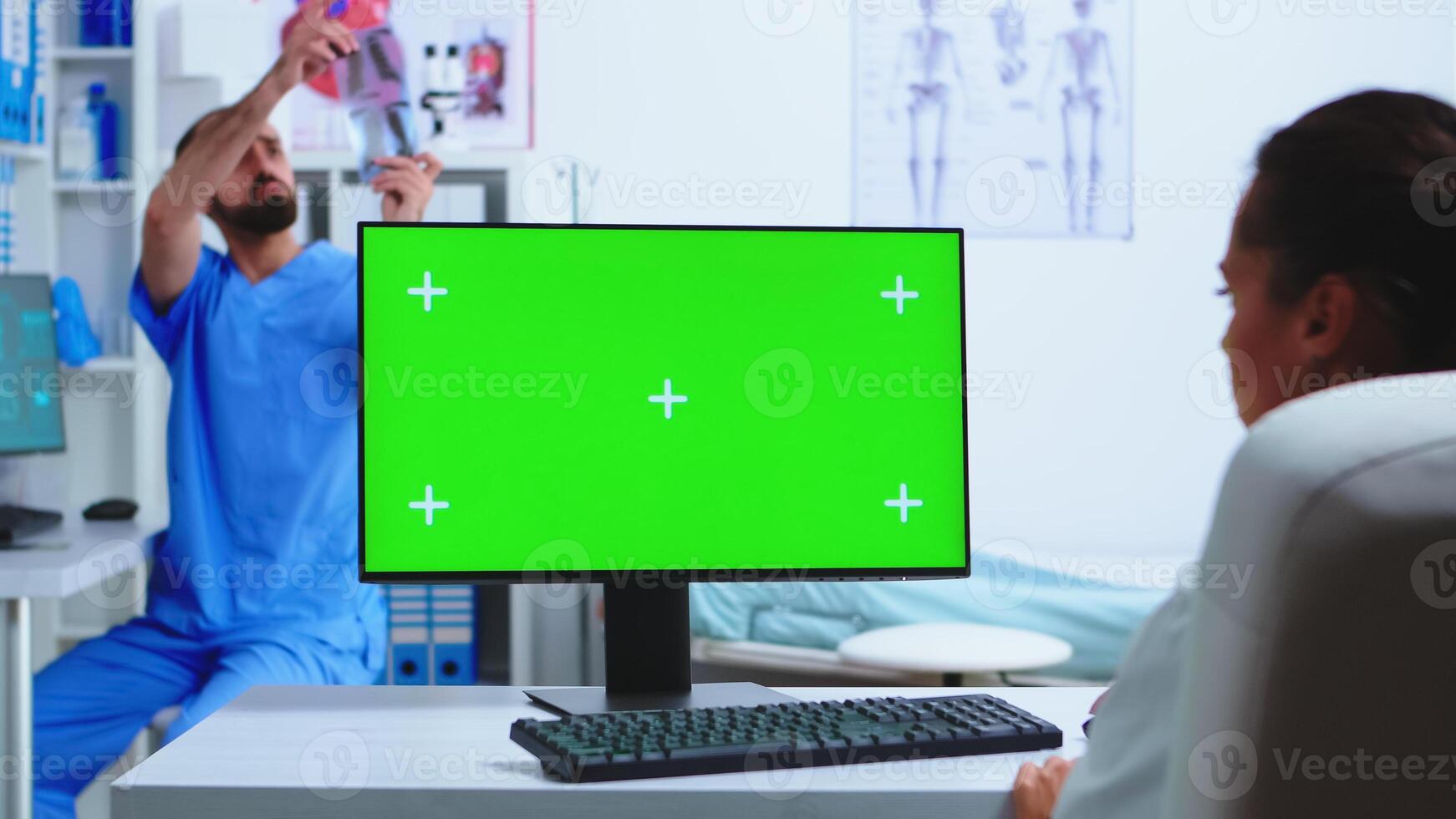 Doctor working on computer with green screen in hospital and assistant looking at x-ray. Desktop with replaceable mockup screen in medical clinic while doctor is checking patient radiography for diagnose. photo