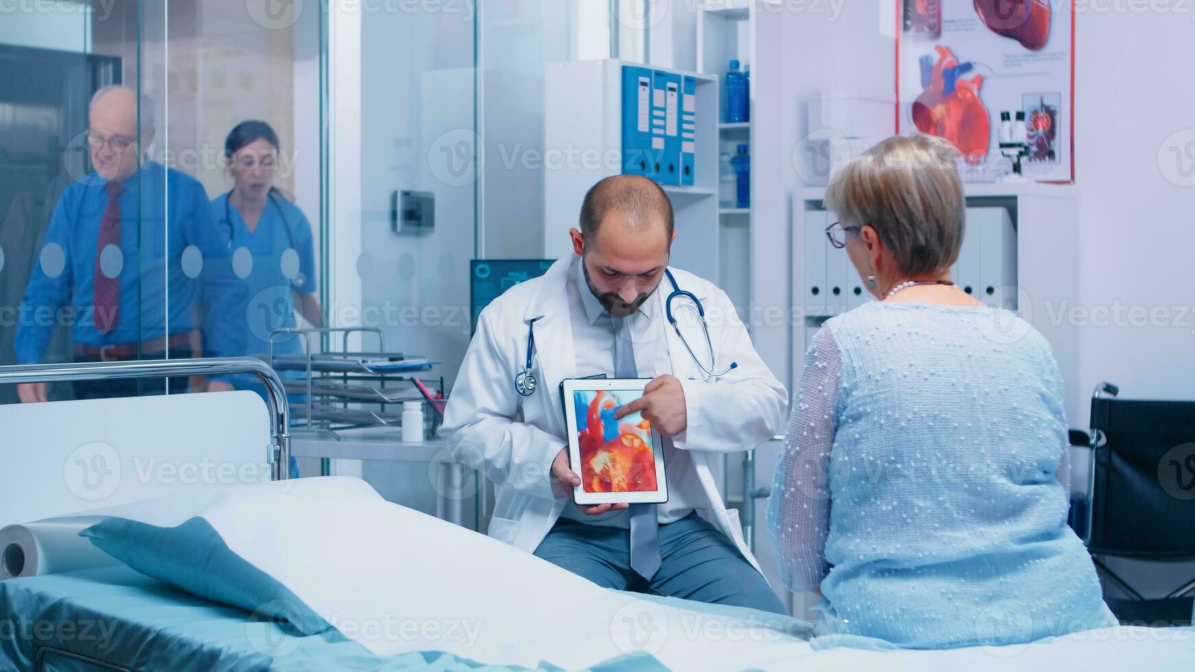 Cardiologyst explaining arrhythmias disease on digital tablet to senior old retired woman sitting on hospital bed in private clinic. Medical medicinal healthcare heart disease diagnostic consultation photo