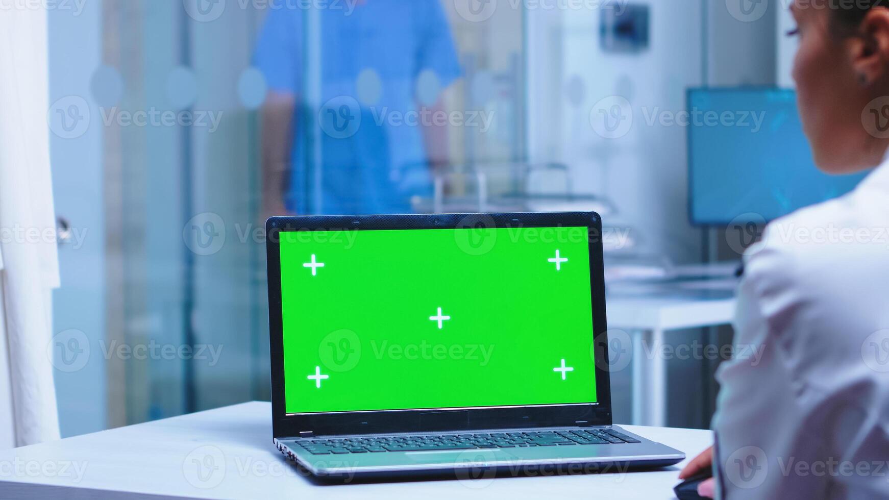 Over shoulder footage of doctor using laptop with copy space available in hospital . Nurse in uniform arriving in cabinet. Medic wearing uniform using notebook with chroma key on display in medical clinic. photo