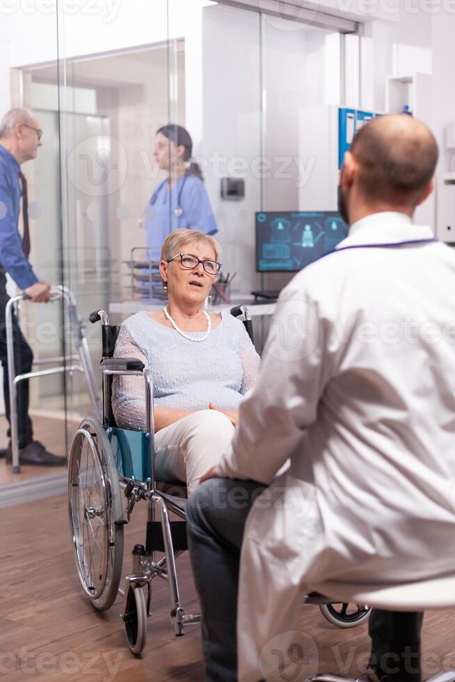 Paralyzed discussing with geriatrician sitting in wheelchair in the course of examination. Medical specialist wearing stethoscope discussing with invalid elderly woman in consultation room of private. photo