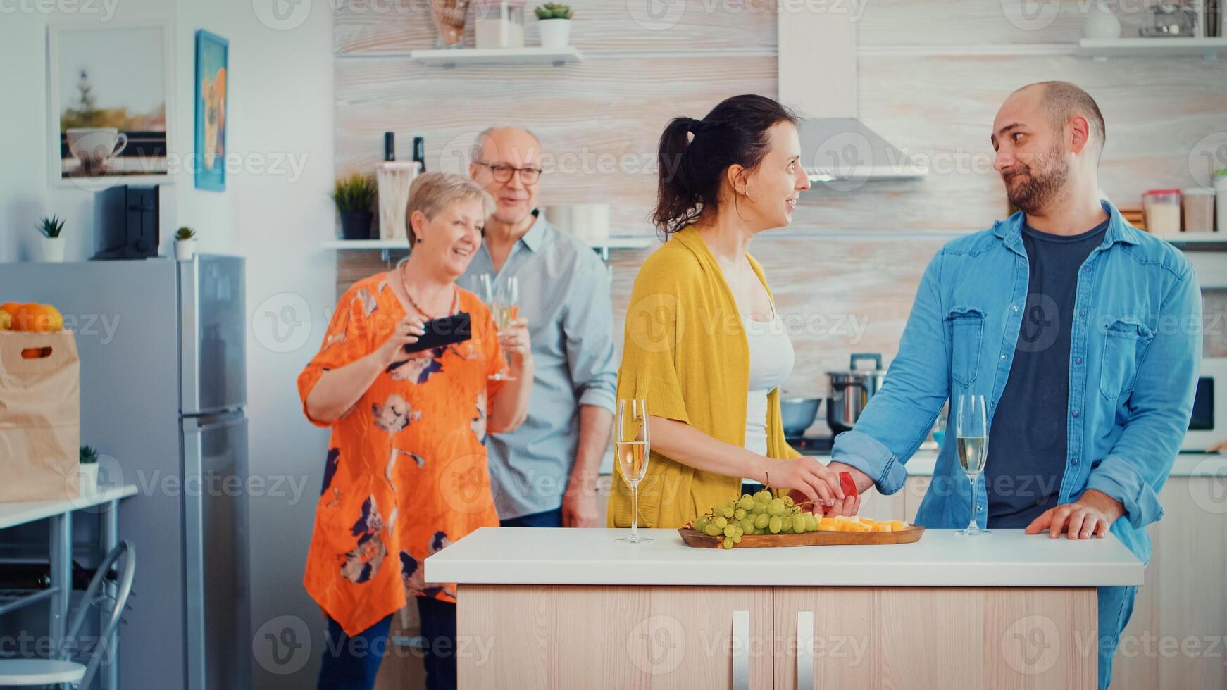 Young man making proposal to his girlfriend in front parents while they talking in the kitchen at home on family day. She putting ring on her finger, kissing and hugging him, mother take pictures photo