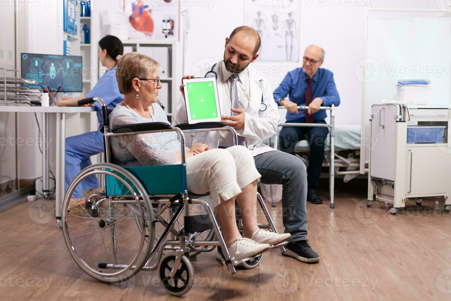 Hospital staff consulting handicapped senior woman sitting in wheelchair holding tablet pc with green screen. Medical practitioner using device with chroma key during coonsultation of invalid senior woman. photo