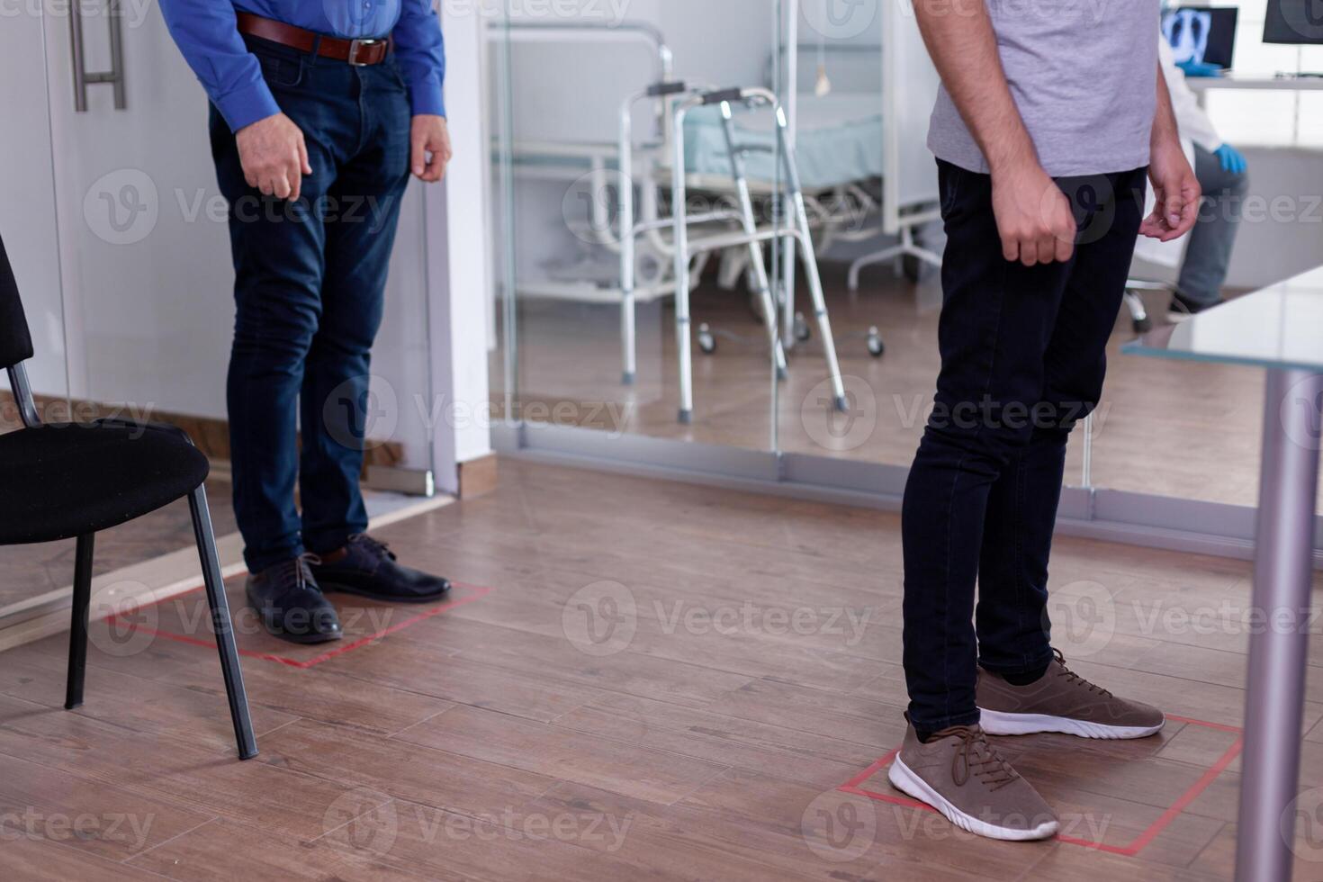 Close up of patients standing in waiting area on floor signs respecting social distancing against coronavirus. Men visiting hospital clinic during covid 19 global pandemic checking appointments. photo