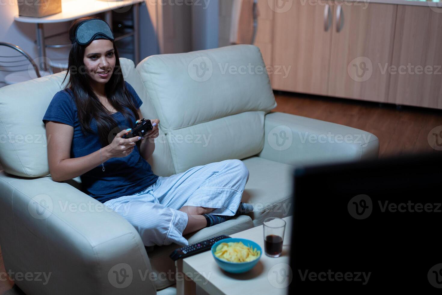 Woman sitting on sofa playing video game late at night wering eye mask on forehead. Excited determined gamer using controller joysticks keypad playstation gaming and having fun winning electronic game photo
