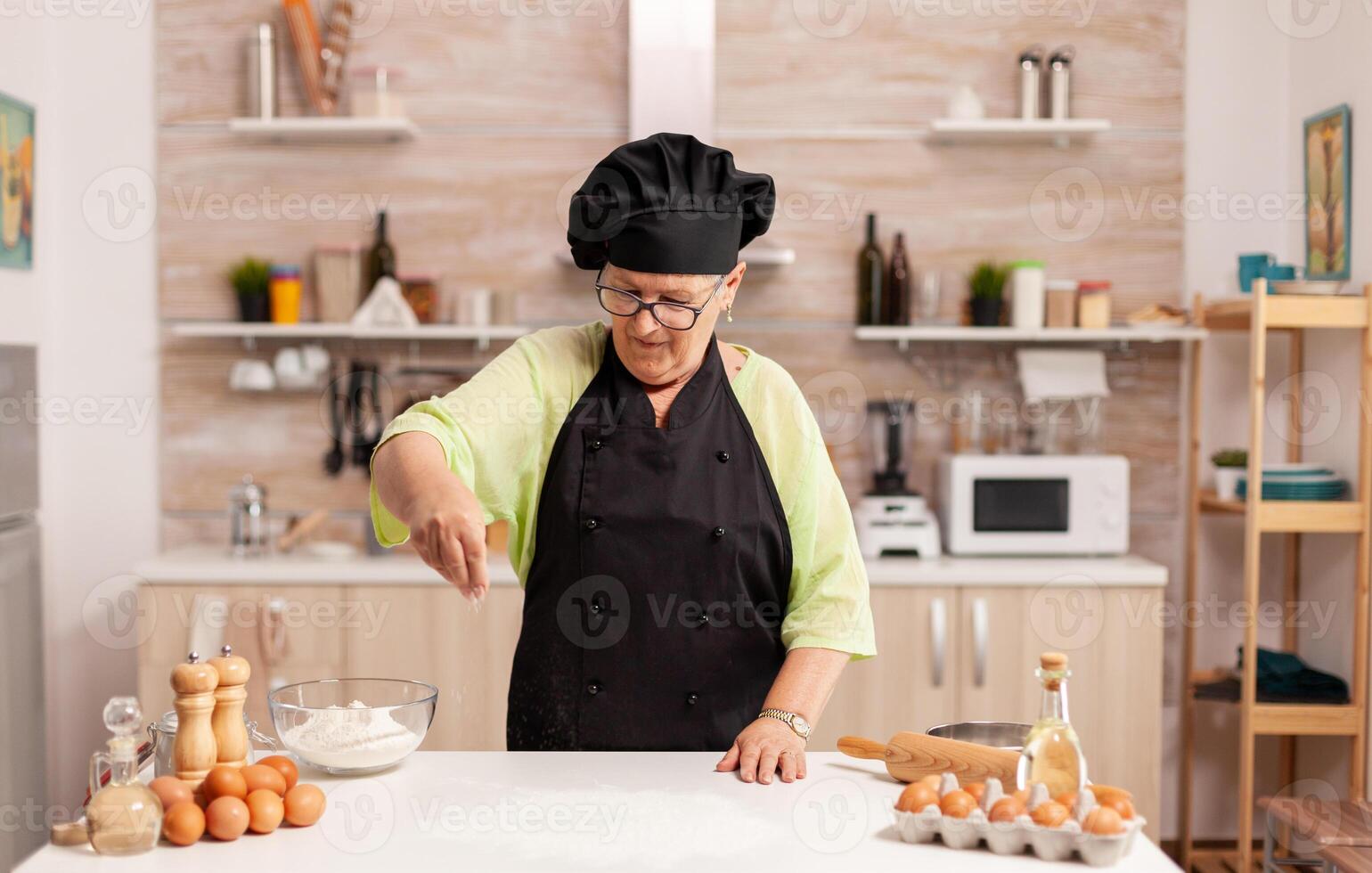 Woman preparing tasty pizza sprinkling baking powder on kitchen table. Happy elderly chef with uniform sprinkling, sieving sifting raw ingredients by hand. photo