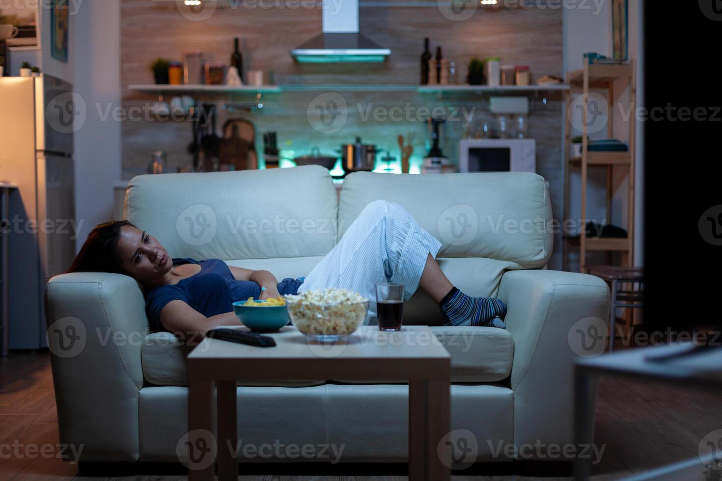 Young woman watching TV and feeling bored sitting on the couch in the living room at home. Tired, exhausted, lonely lady relaxing watching tv lying on comfortable sofa eating snacks late at night photo