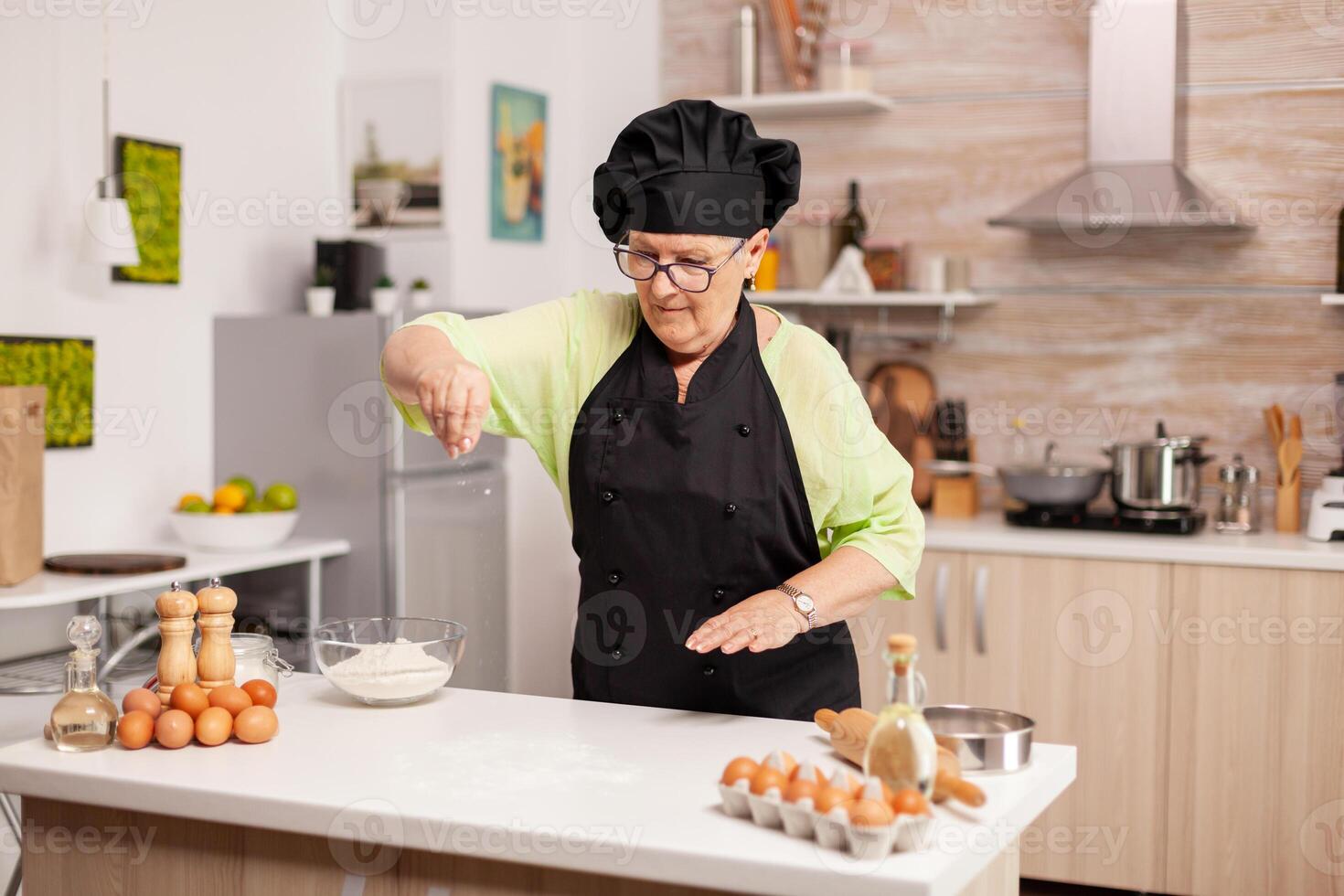 Retired chef preparing pizza in home kitchen wearing apron. Happy elderly chef with uniform sprinkling, sieving sifting raw ingredients by hand in home kitchen. photo