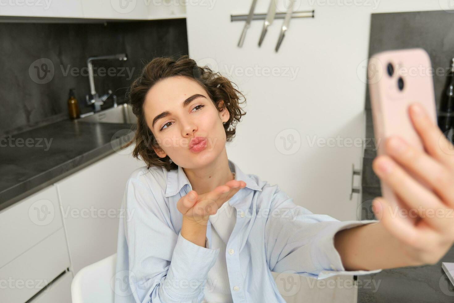 Portrait of cute brunette woman takes selfie, sends kisses at smartphone camera, records video on mobile phone app photo