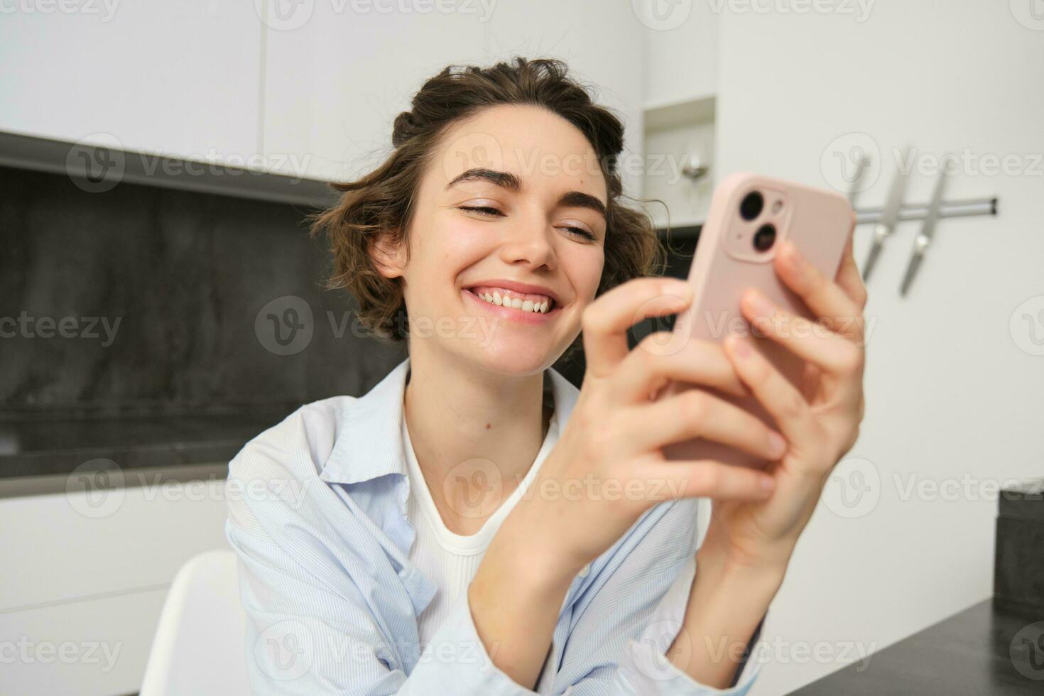Portrait of brunette woman chats on mobile phone, uses smartphone app from home, orders delivery while sits in kitchen photo