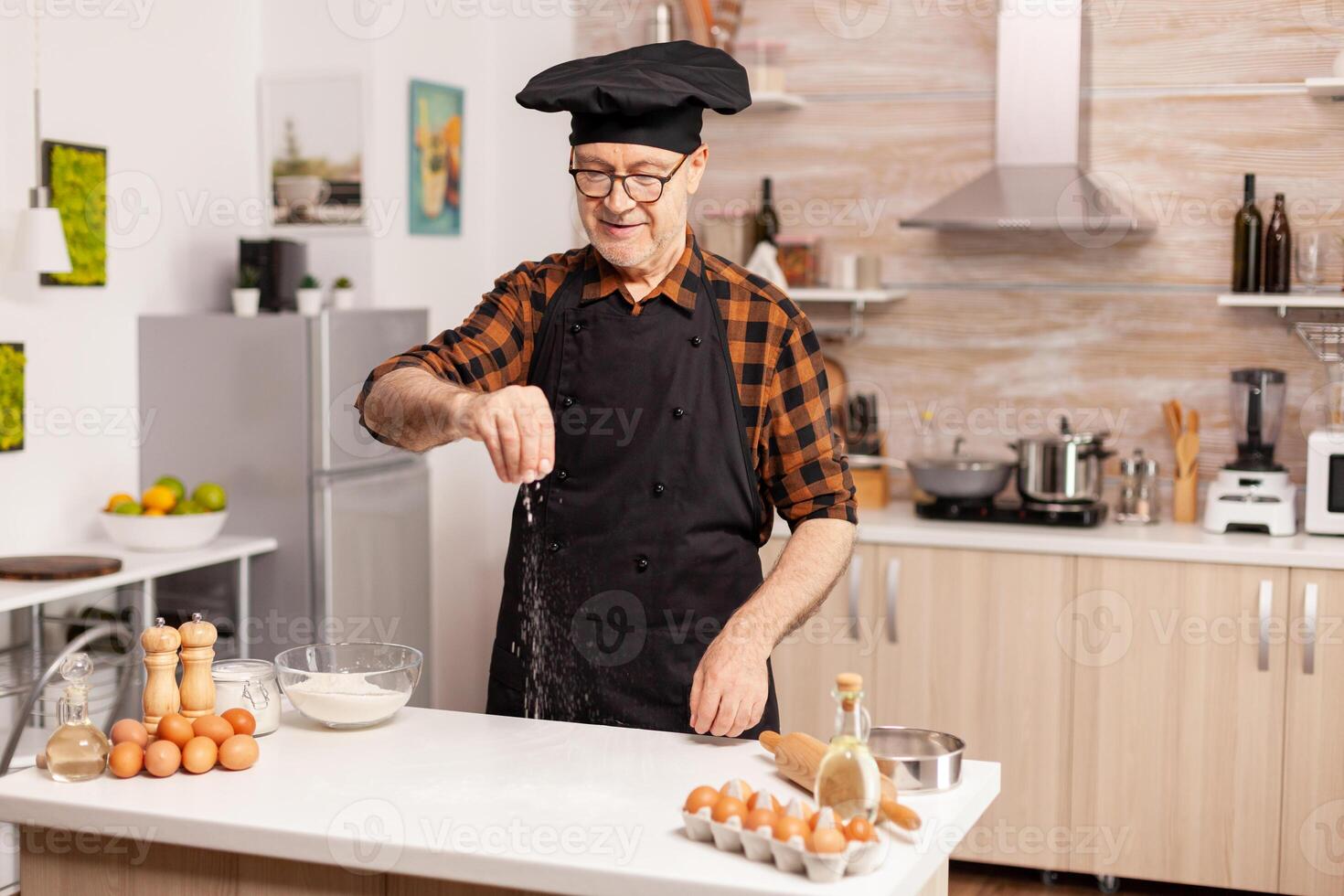 Experienced baker in kitchen preparing delicious pizza using bio wheat flour. Retired senior chef with bonete and apron, in kitchen uniform sprinkling sieving sifting ingredients by hand. photo