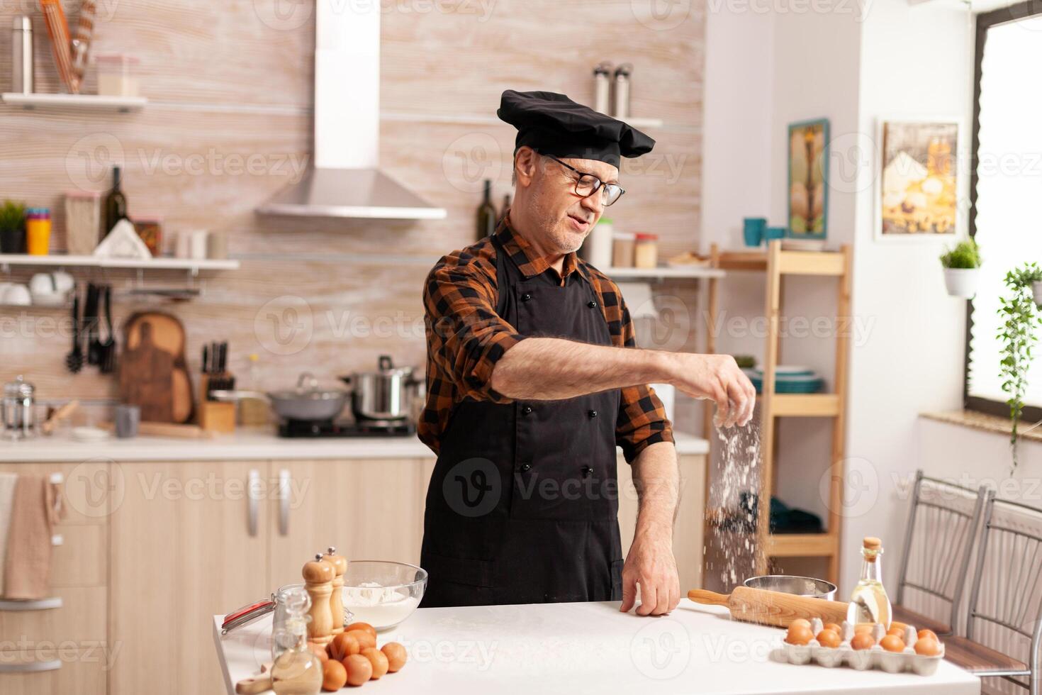 Chef in home kitchen gluten free dough for pasta, bakery or pizza. Retired senior chef with bonete and apron, in kitchen uniform sprinkling sieving sifting ingredients by hand. photo