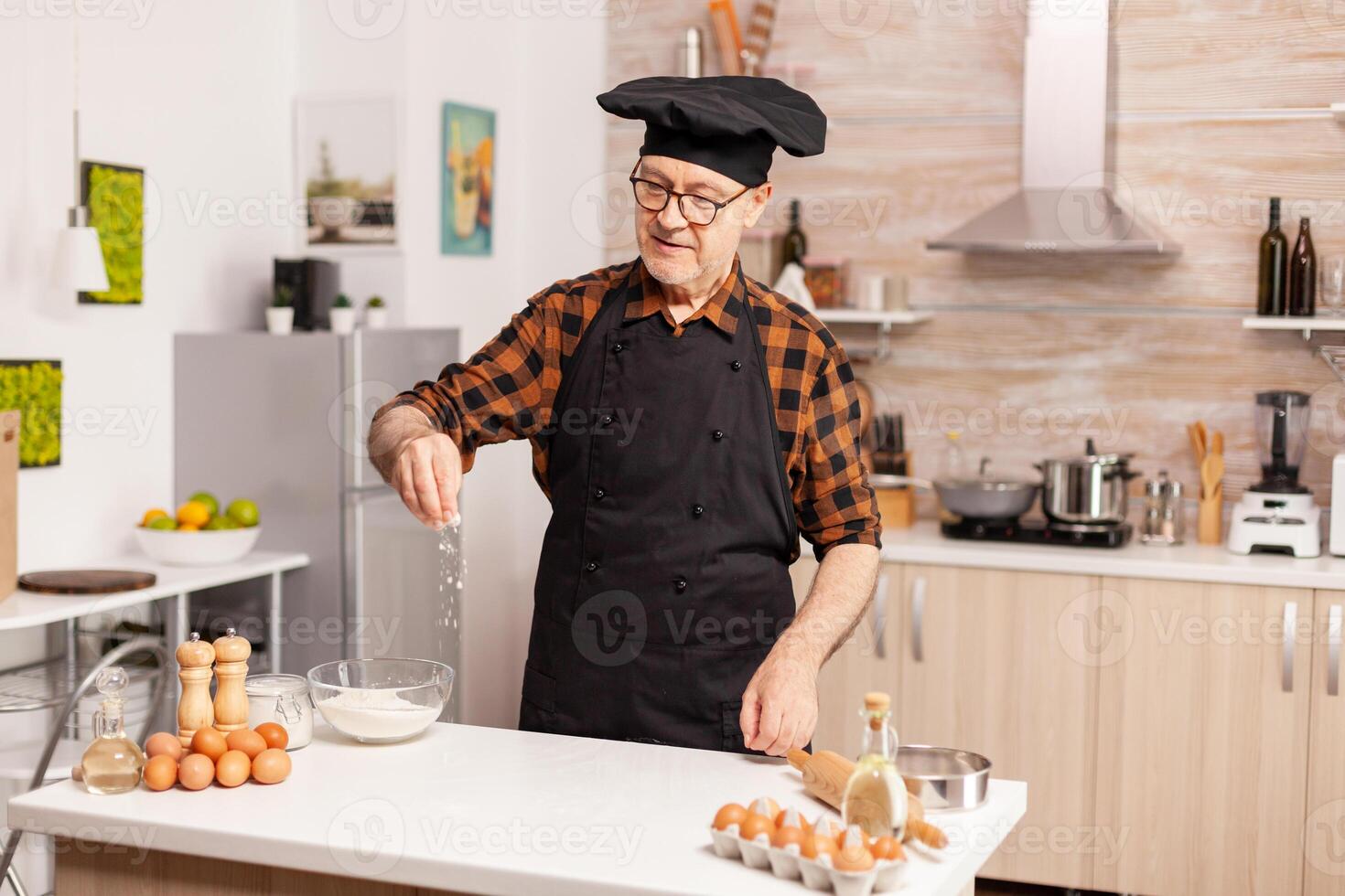 Senior baker in home kitchen putting wheat flour over wooden table. Retired senior chef with bonete and apron, in kitchen uniform sprinkling sieving sifting ingredients by hand. photo
