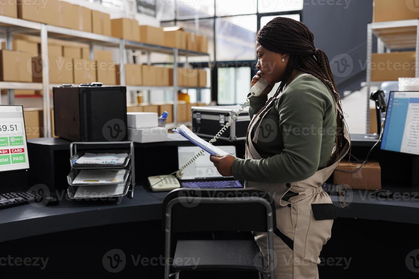 Stockroom african american worker holding clipboard, analyzing goods inventory report in storehouse. Storage room employee talking at landline phone with remote customer discussing delivery details photo