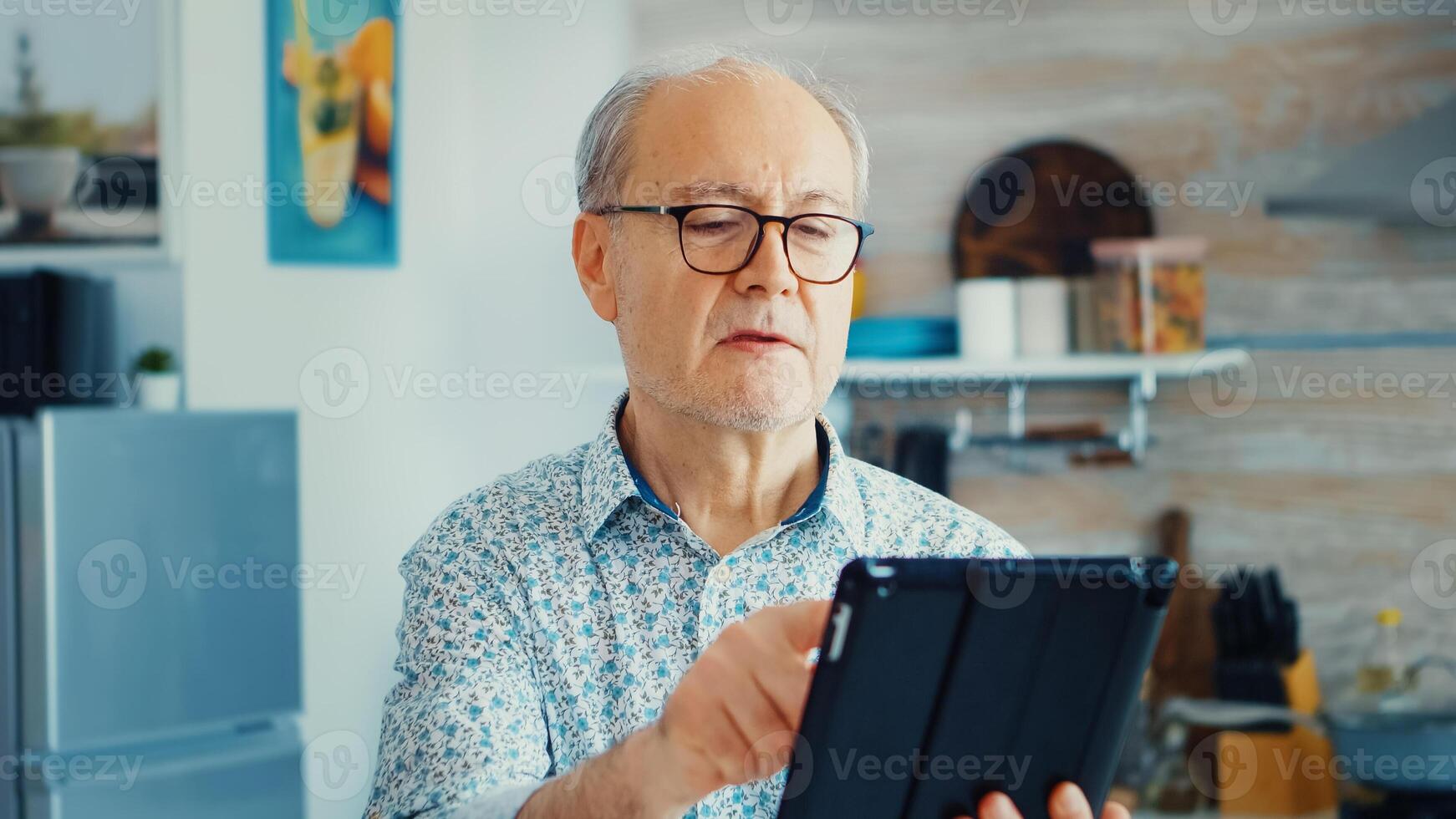 Elderly man using tablet pc in kitchen during breakfast wearing glasses. Elderly person with tablet portable pad PC in retirement age using mobile apps, modern internet online information technology with touchscreen photo