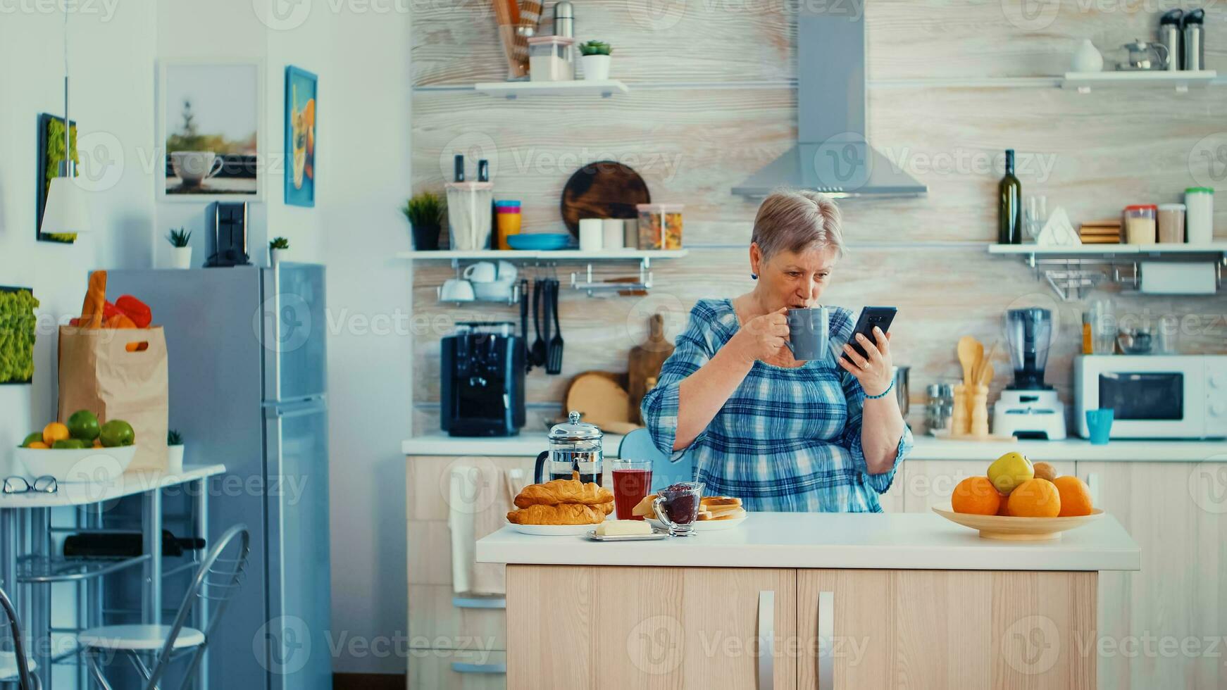 mayor mujer diciendo Hola mientras teniendo un vídeo llamada con familia utilizando teléfono inteligente en cocina durante desayuno. mayor persona utilizando Internet en línea charla tecnología vídeo cámara web haciendo un vídeo llamada conexión cámara comunicación conferencia llamada foto