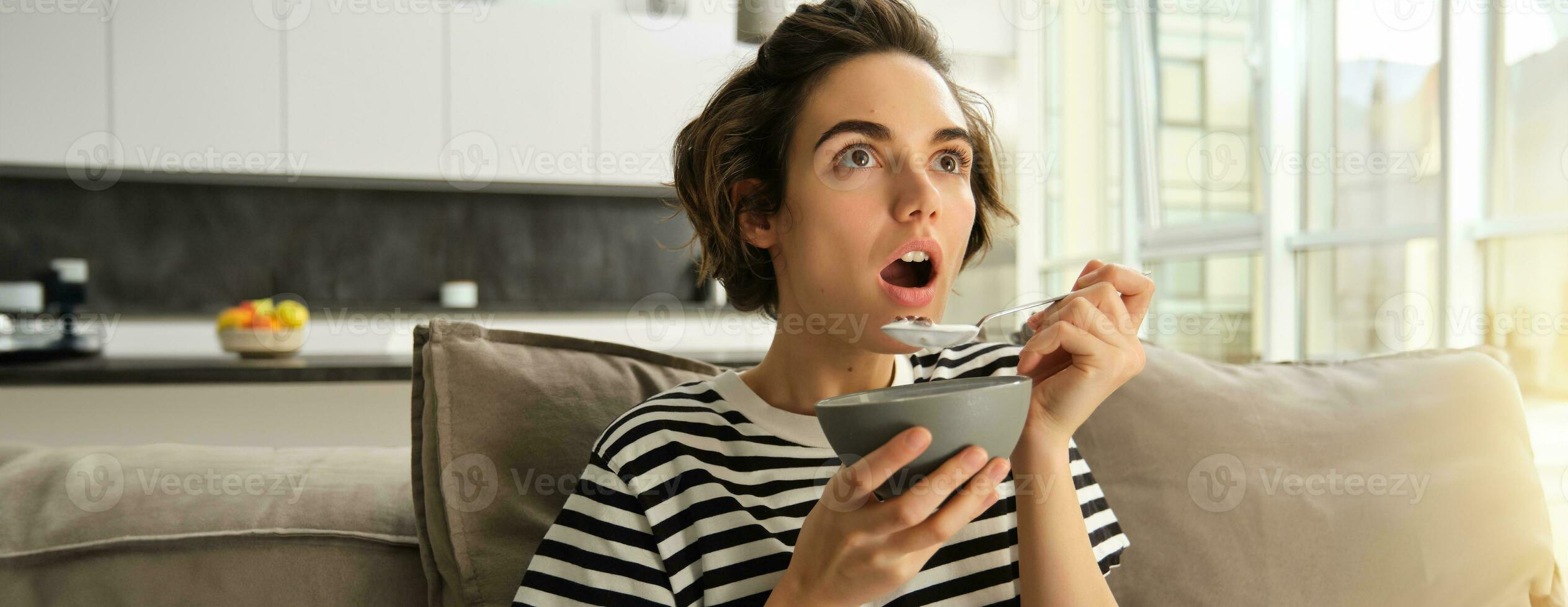 Close up portrait of woman eating in front of tv, looking amazed and with interest at screen, eating breakfast, holding bowl of cereals and spoon, sitting on sofa fascinated photo
