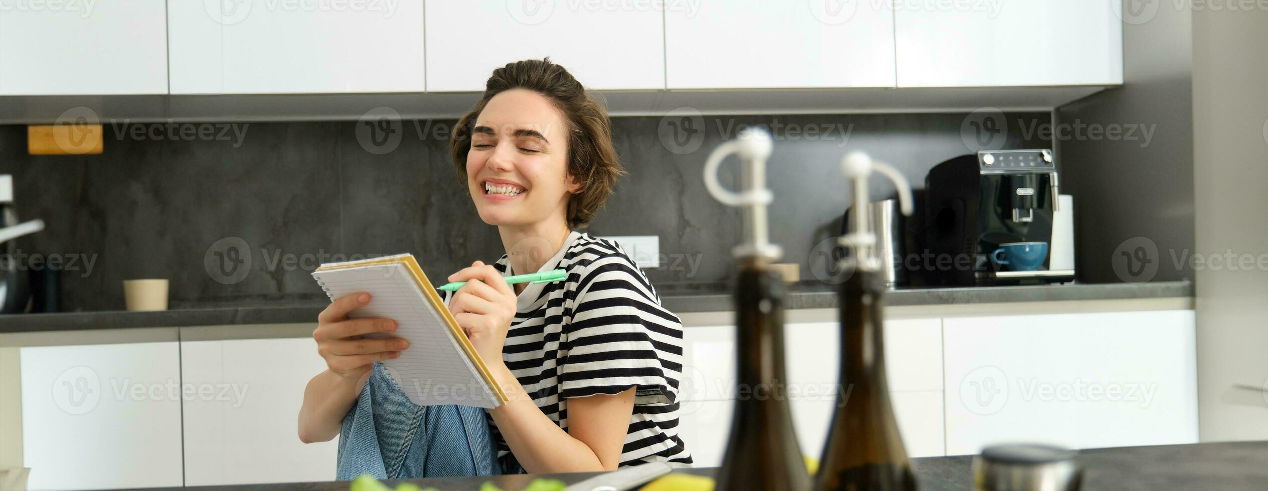 Close up of happy, smiling young woman writing in notebook, creating recipe, list of meals for this week, sitting near vegetables and olive oil, cooking in the kitchen photo