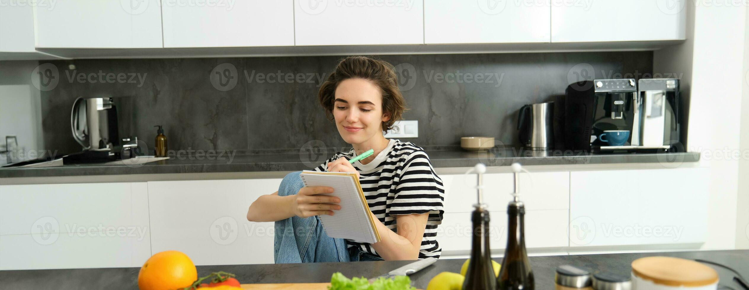 Smiling brunette woman in kitchen, writing down meal list for week, reading recipe, cooking salad, vegetable meal at home photo