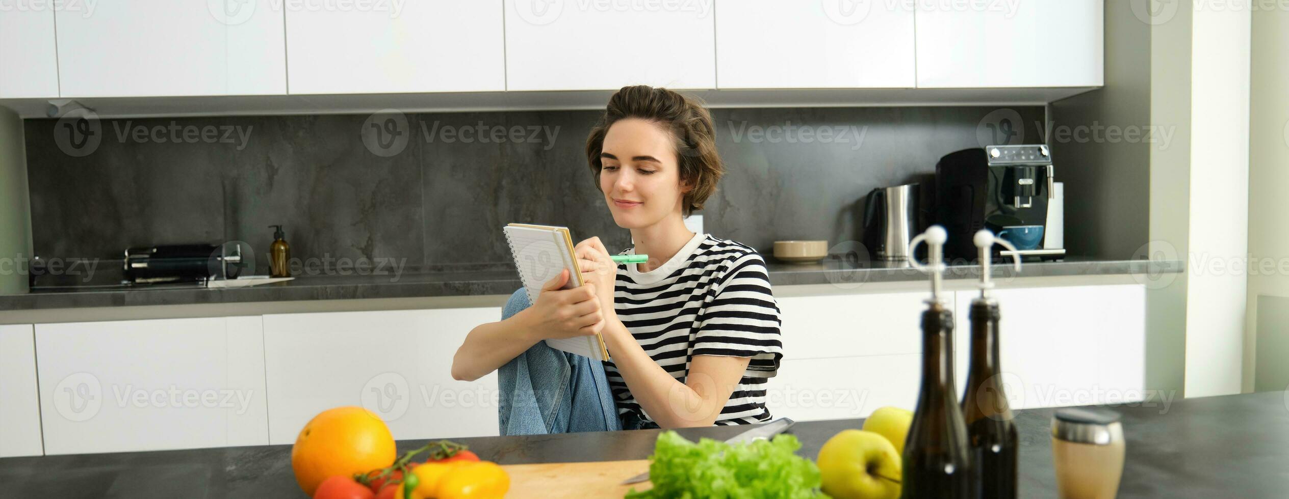 Portrait of beautiful modern woman, girl cooking with vegetables, holding notebook, writing down recipe, making notes, dinner plans, creating meal list for week, sitting in kitchen photo