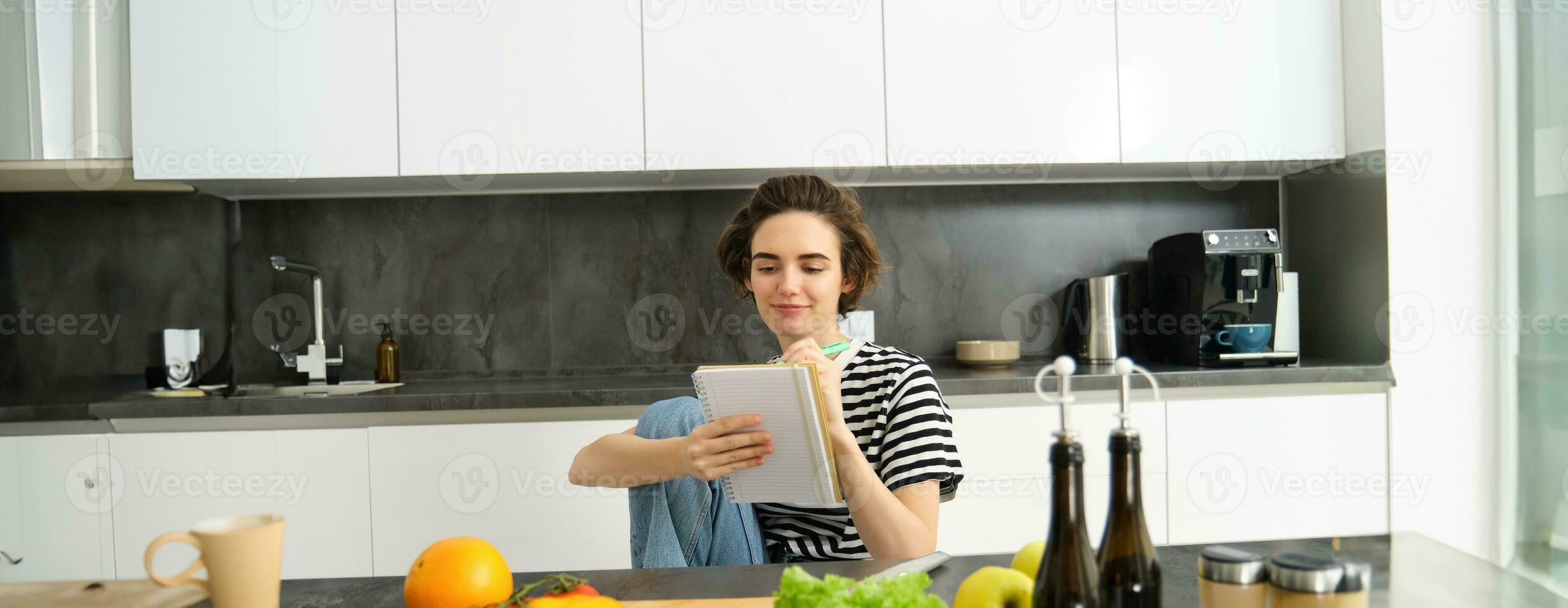 Portrait of young smiling woman cooking with vegetables, writing down ingredients, looking at her notebook, reading recipe, sitting in kitchen photo