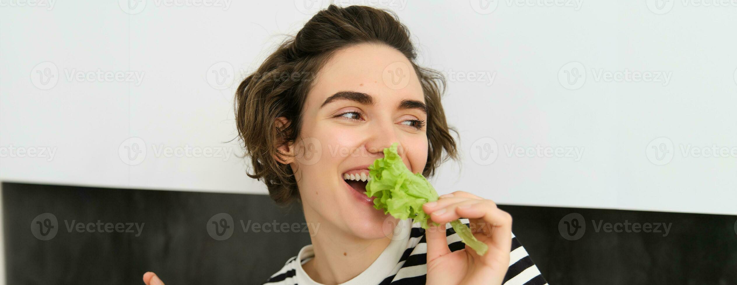 retrato de despreocupado vegetariano chica, comiendo verduras, mordedura lechuga hoja con contento sonriente rostro, teniendo sano bocadillo, gustos verduras, soportes en el cocina foto