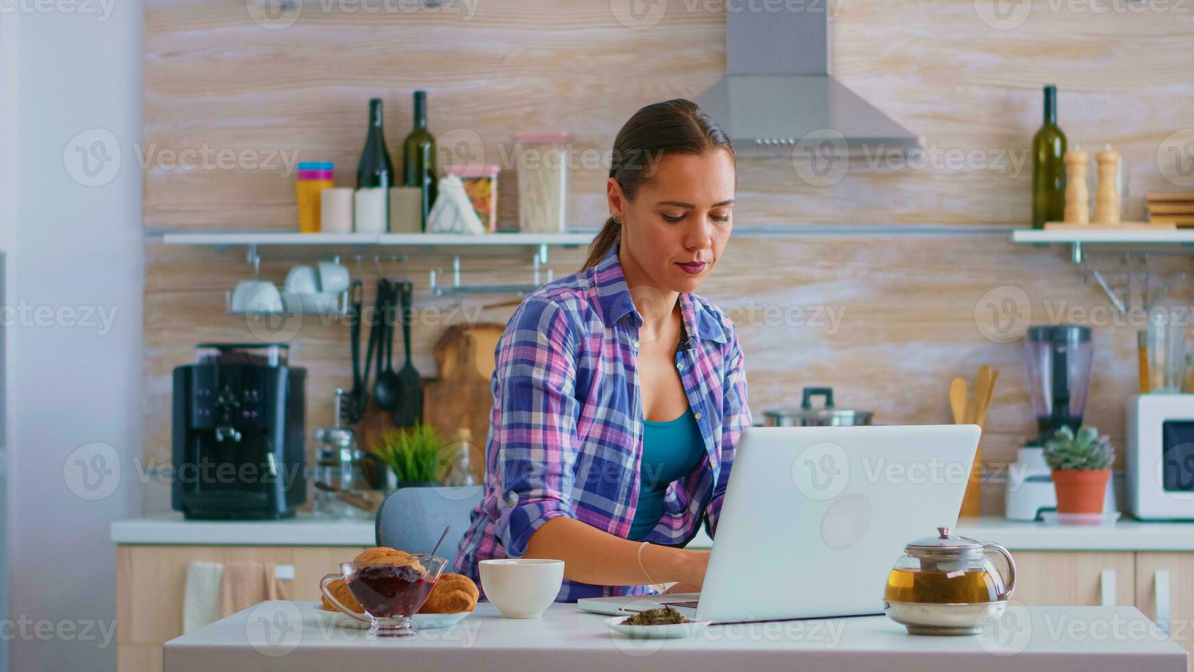 Woman sipping green tea and typing on her laptop computer during breakfast in cozy kitchen. Working from home using device with internet technology, browsing, searching on gadget in the morning. photo