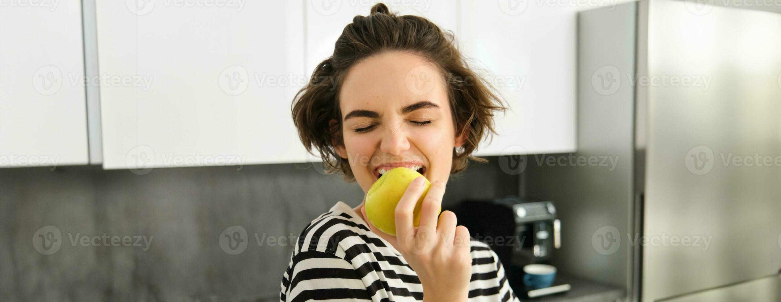 Close up portrait of young brunette woman biting an apple with pleasure, has pleased smile on her face, standing in the kitchen, having healthy snack for lunch, eating fruits photo