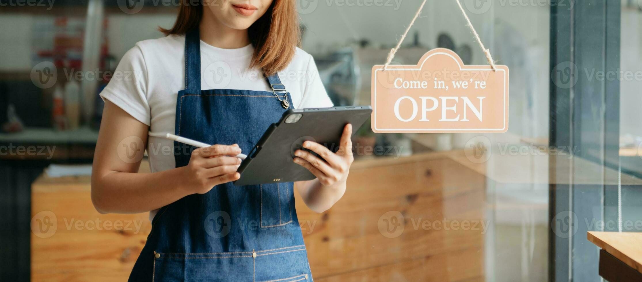 Young female hanging a welcome sign in front of a coffee shop. Beautiful waitress or hostess holding a tablet preparing  in a restaurant. photo