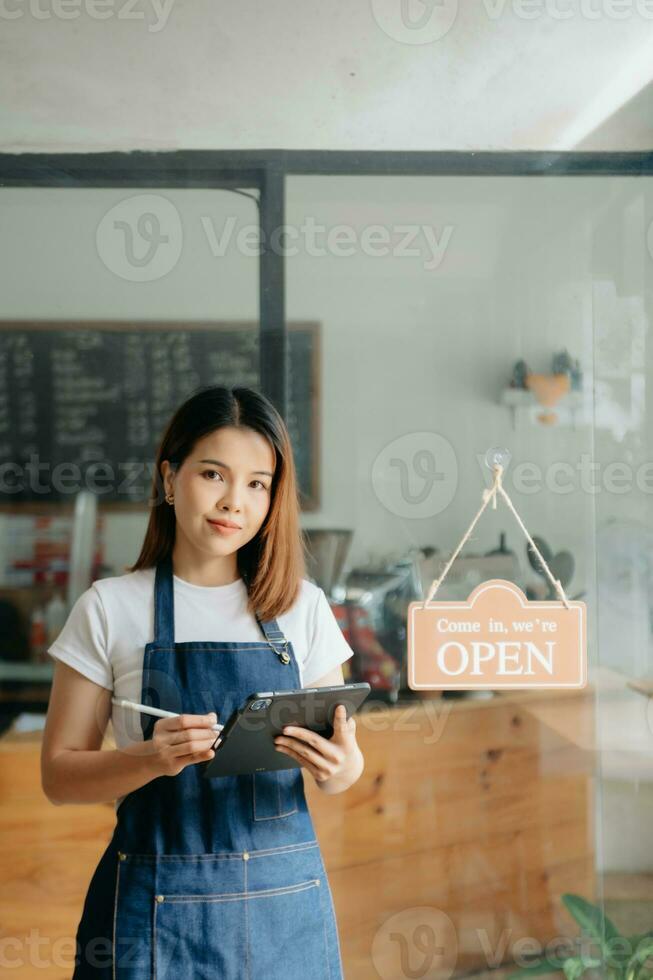 joven hembra colgando un Bienvenido firmar en frente de un café tienda. hermosa camarera o anfitriona participación un tableta preparando en un restaurante. foto