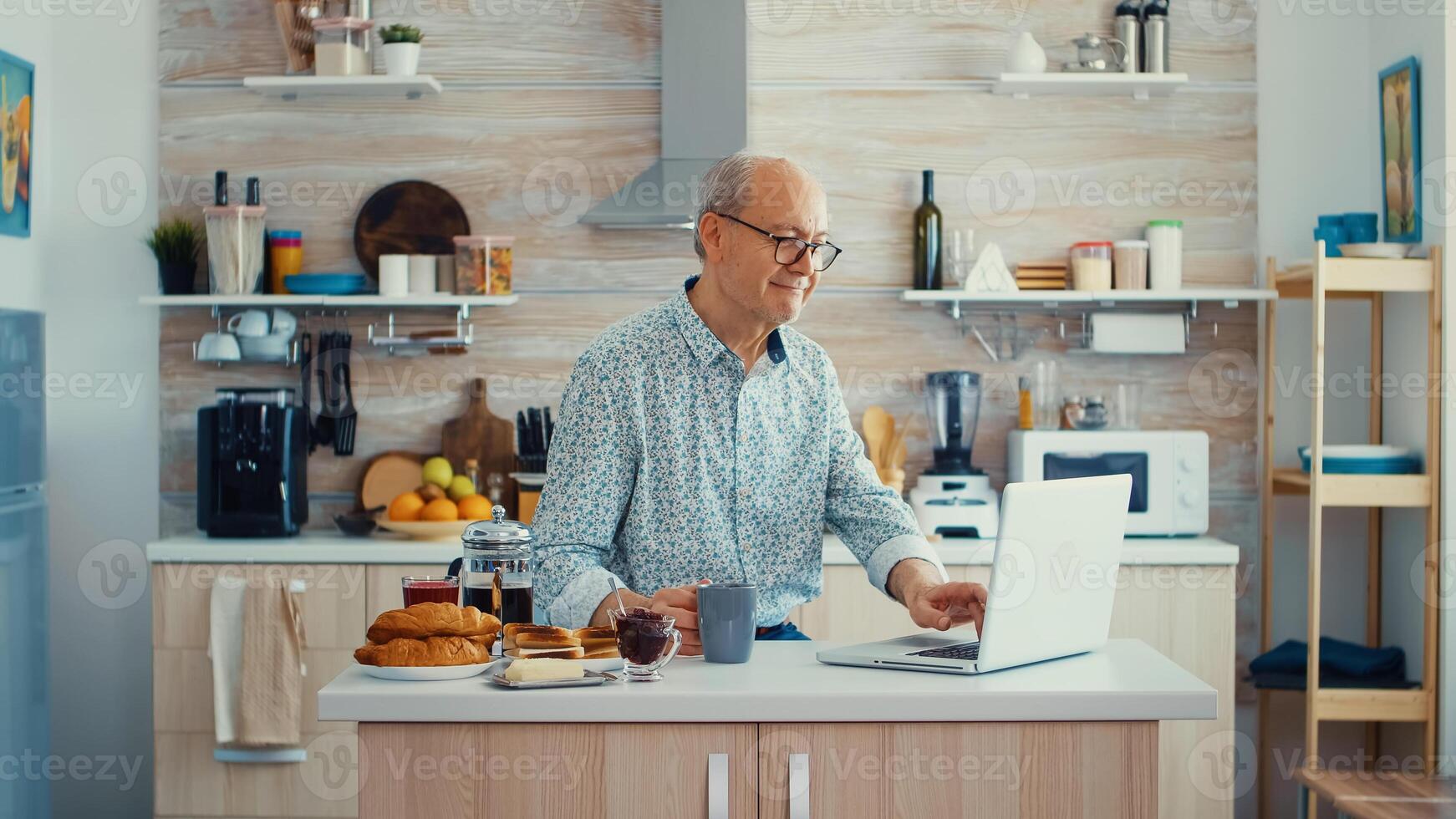 Daily life of senior man in kitchen during breakfast using laptop holding a cup of coffee. Elderly retired person working from home, telecommuting using remote internet job online communication on modern technology notebook photo