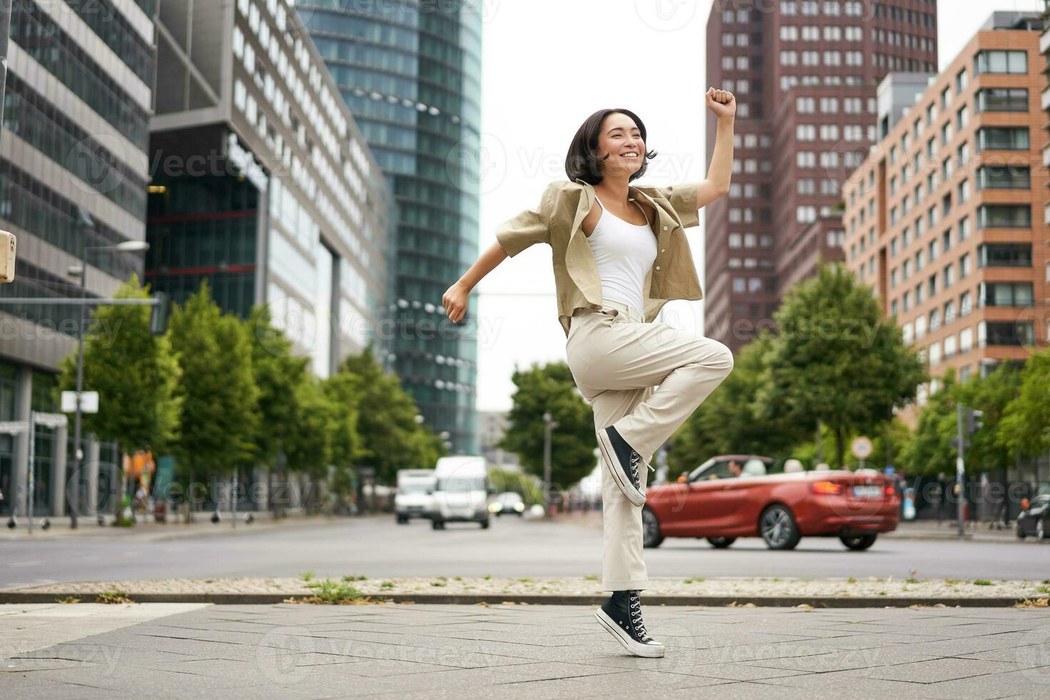 Portrait of asian happy girl jumping and dancing in city centre, posing on streets, express joy and excitement photo