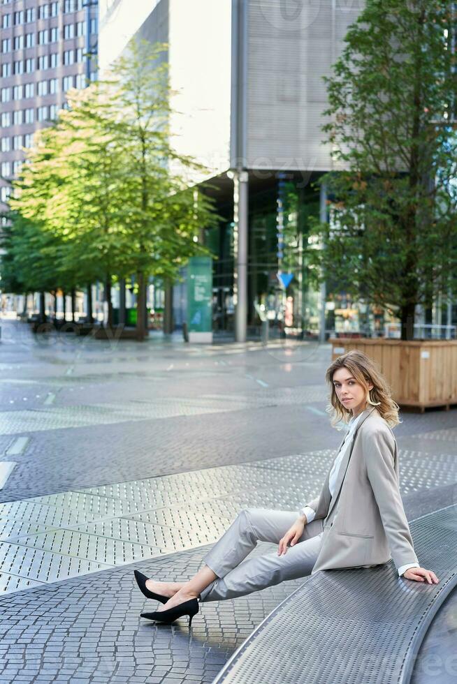 Vertical shot of beautiful businesswoman in beige suit, sitting outdoors in city centre and smiling at camera photo