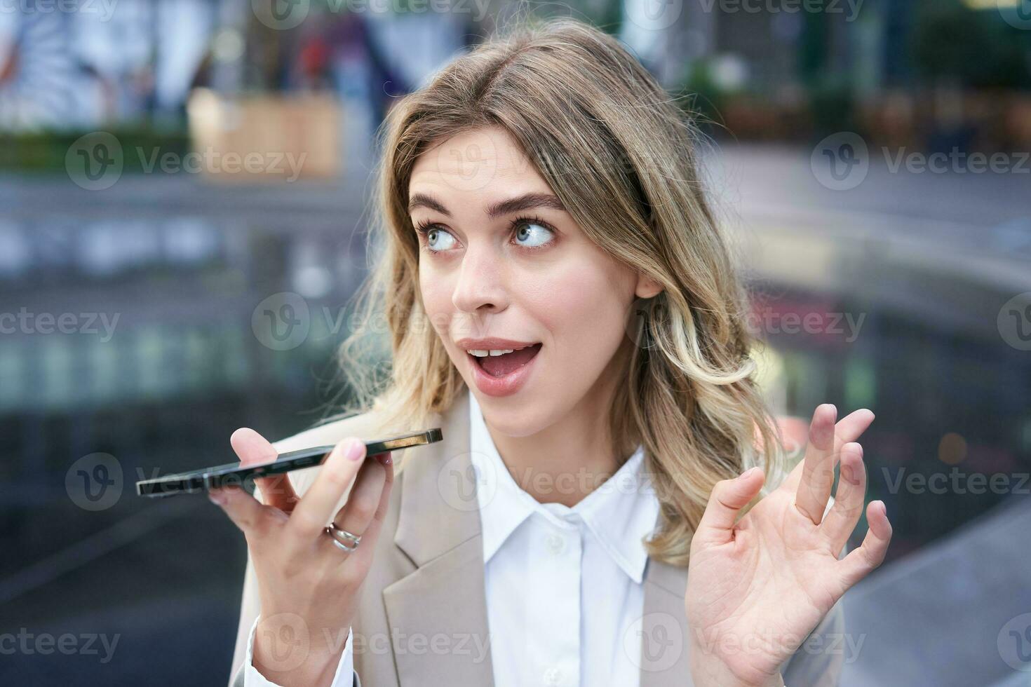 Chatty businesswoman sitting in city centre and recording voice message, talking into microphone on smartphone photo