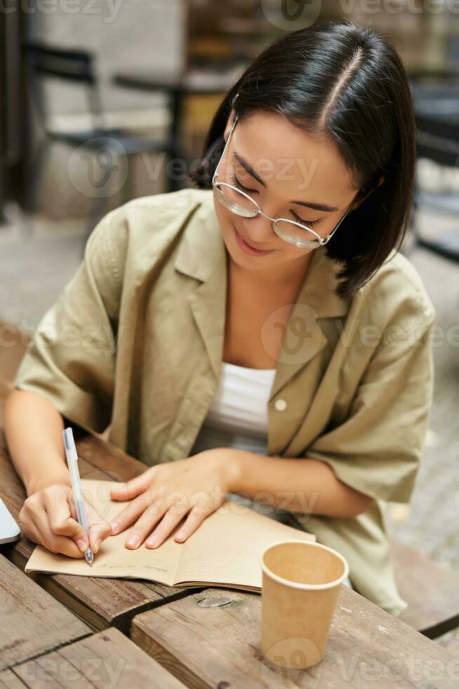 Vertical shot of young asian woman doing homework, making notes, writing something down, sitting in an outdoors cafe and drinking coffee photo
