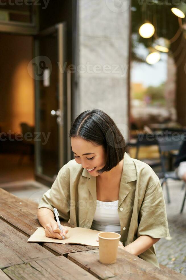 Vertical shot of asian girl student, writing smth in notebook, making notes, smiling while doing homework outdoors from cafe photo