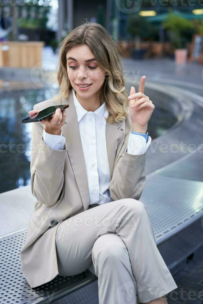 Vertical shot of businesswoman in suit, talking in microphone on phone, record voice message, using speakerphone while sitting on empty street near office buildings photo