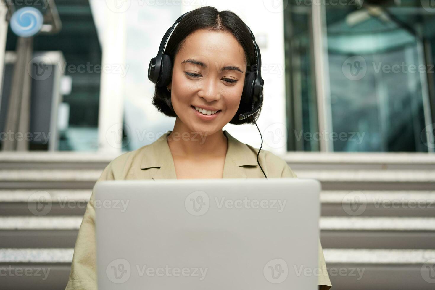 Portrait of young asian woman sitting with laptop and headphones, watching video, does online course on computer, sitting on stairs outdoors photo